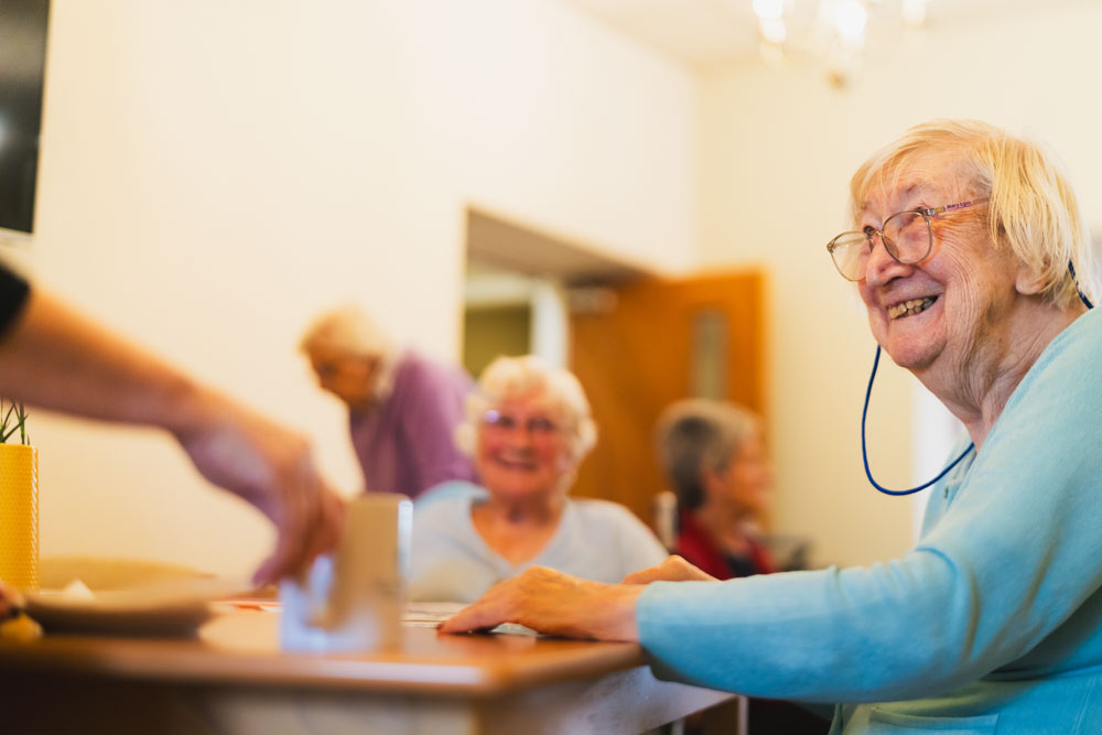 An elderly woman with glasses smiles whilst sitting at a table in a bright, cosy room with other pensioners. Another person's hand reaches towards her, possibly serving or assisting.