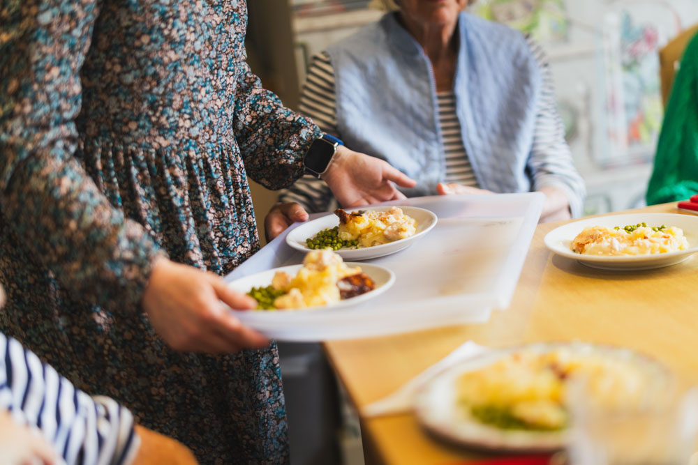 A person serves plates of food on a tray to people sitting at a table, suggesting a meal in a casual or communal dining setting.