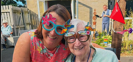 Two smiling women wear large, colourful novelty glasses decorated with flowers and birds whilst posing together outdoors on a sunny day.